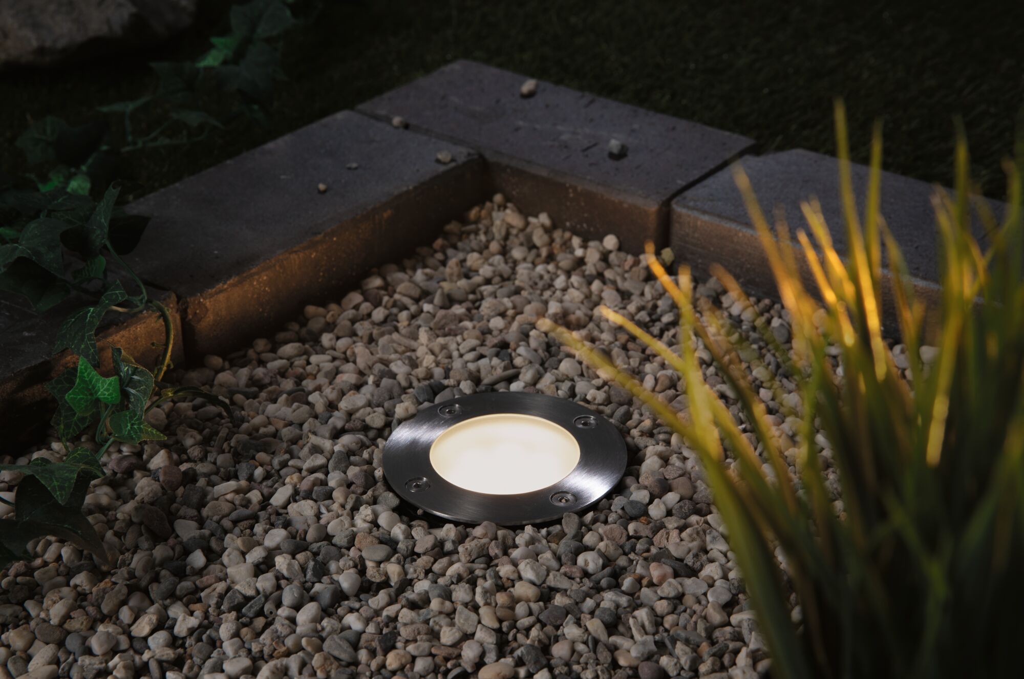 Round stainless steel recessed light with warm white glow in gravel bed, outdoor garden lighting