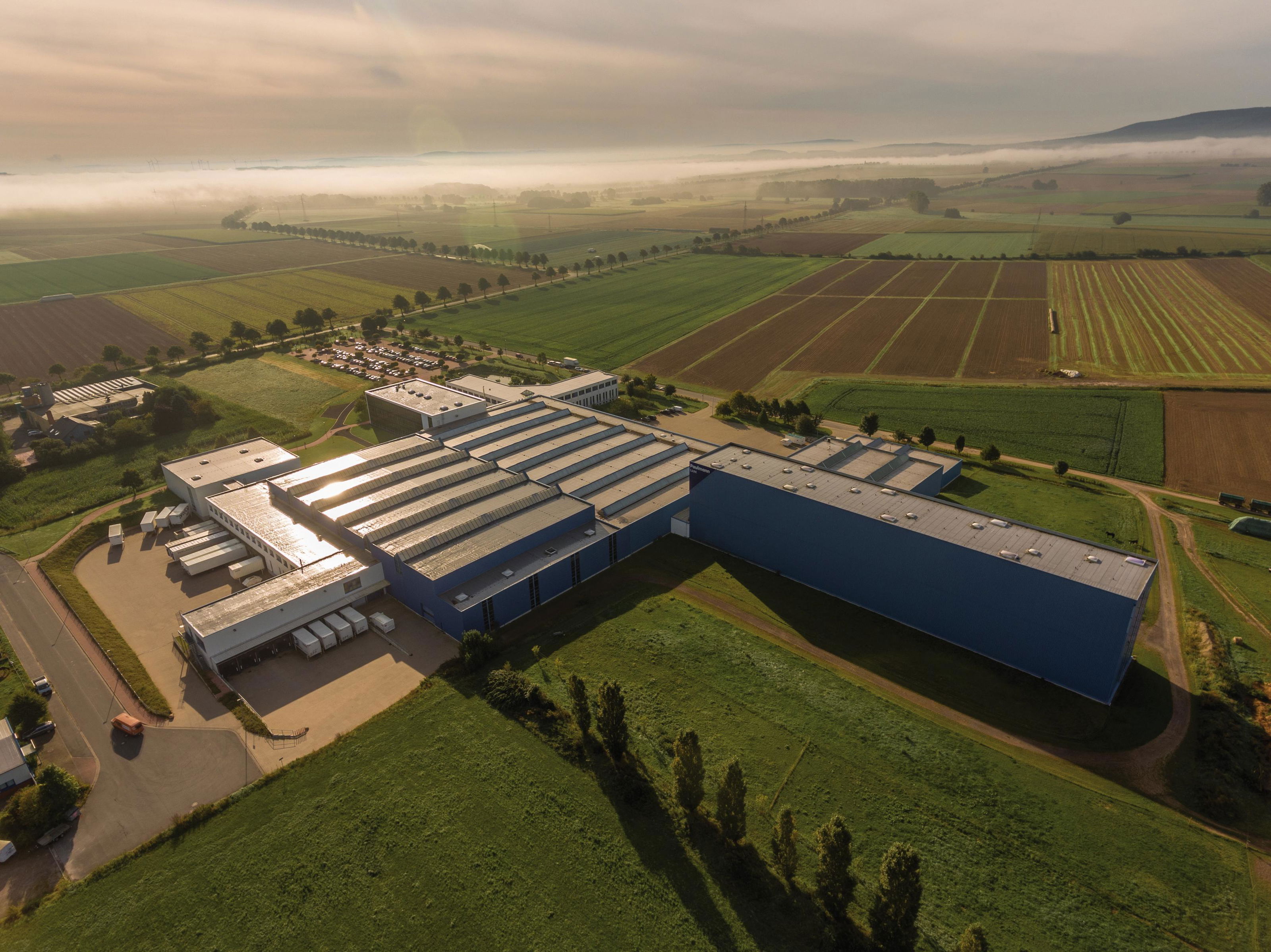 Vue aérienne d’un grand bâtiment industriel bleu entouré de champs verts et parkings au lever du soleil