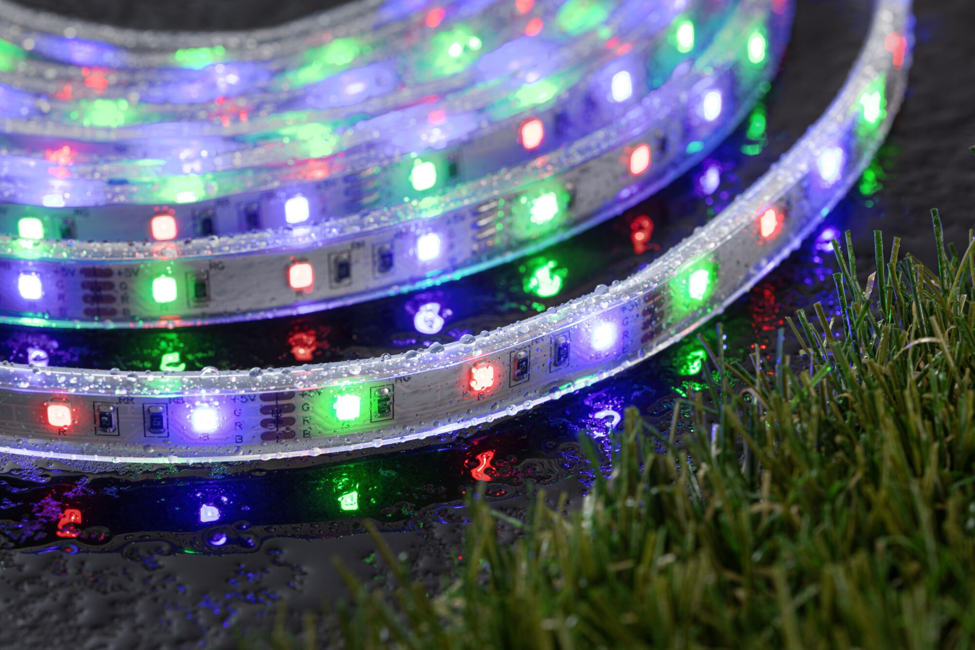 Colorful LED strip light with waterproof plastic casing on wet surface near grass, outdoor lighting.