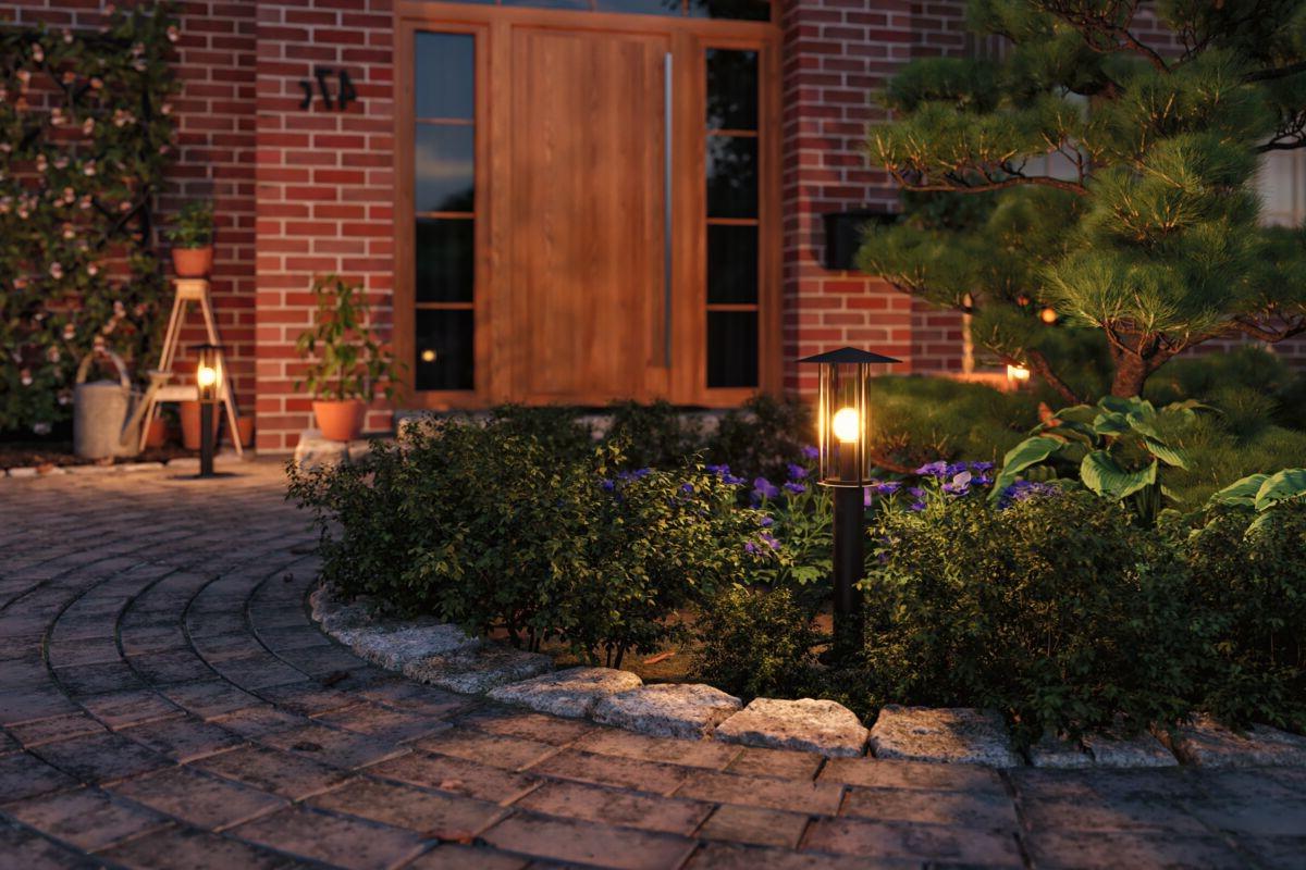 Lampe extérieure noire moderne avec lumière blanche chaude dans jardin devant porte en bois et mur en briques.