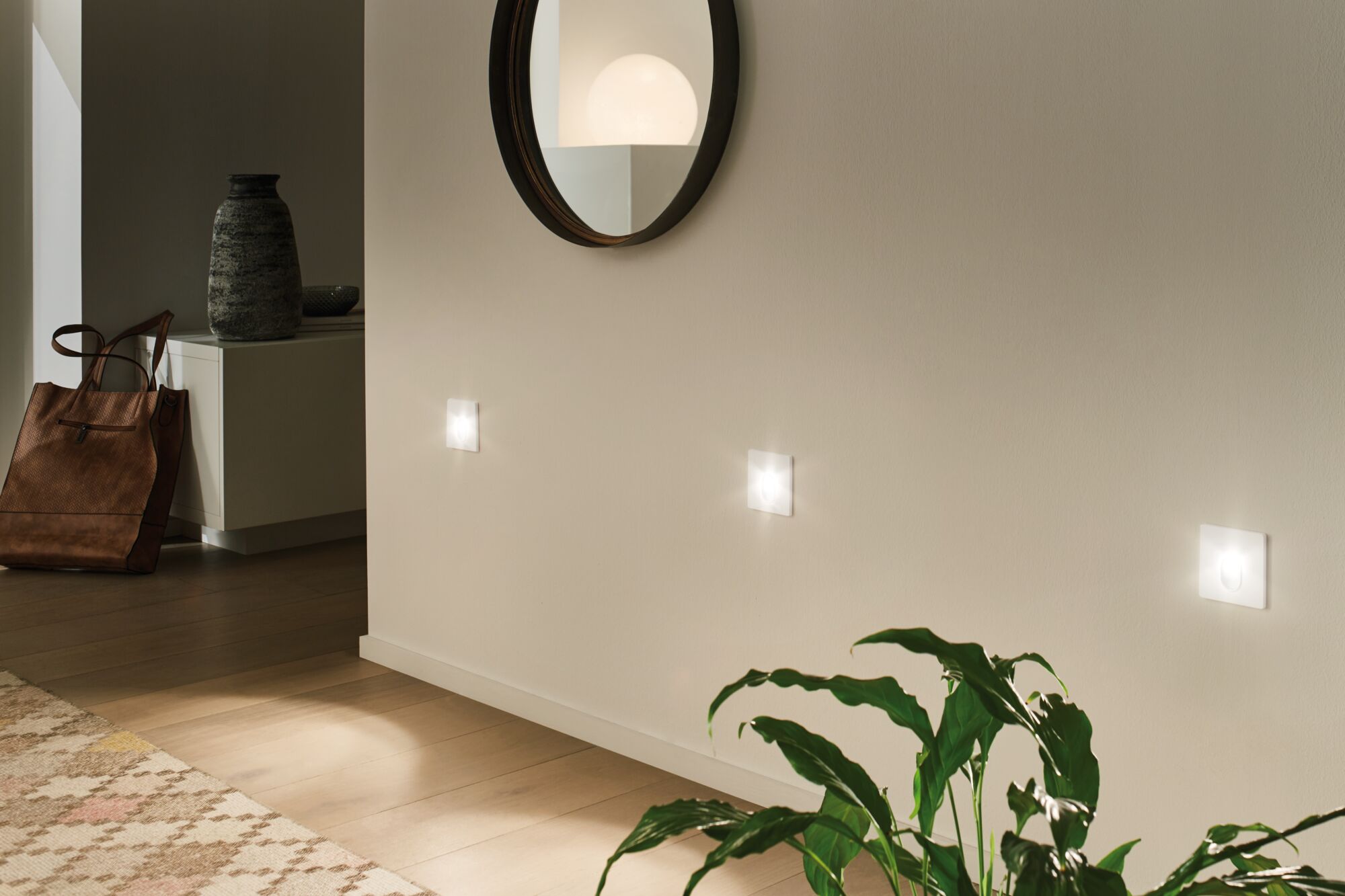 Drie witte LED-wandlampen aan beige muur in moderne hal met spiegel en plant