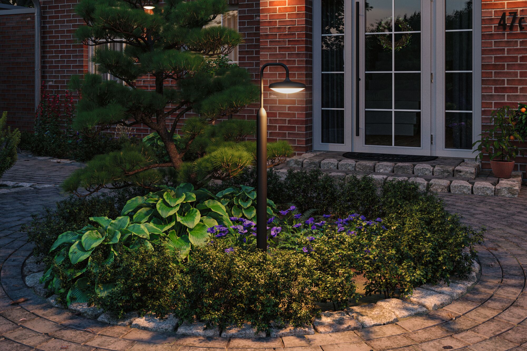 Black outdoor lamp with round shade lighting garden bed in front of brick house with glass door