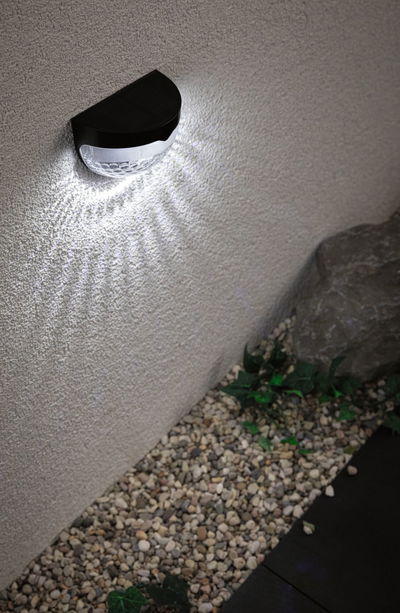 Black solar wall light with white LED on exterior wall above gravel and plants for garden lighting