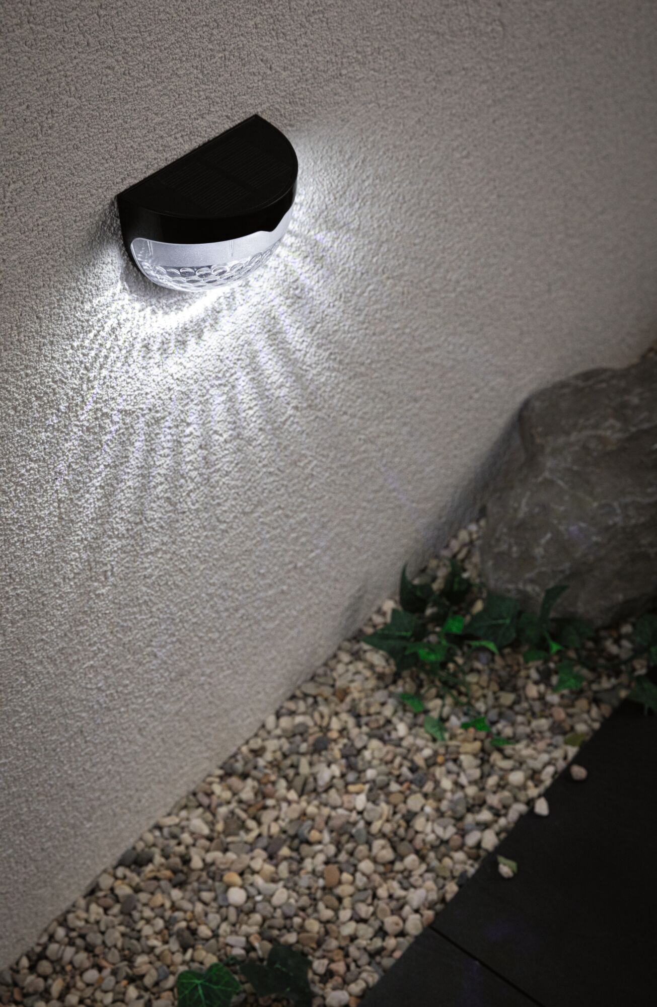 Black solar wall light with white LED on exterior wall above gravel and plants for garden lighting