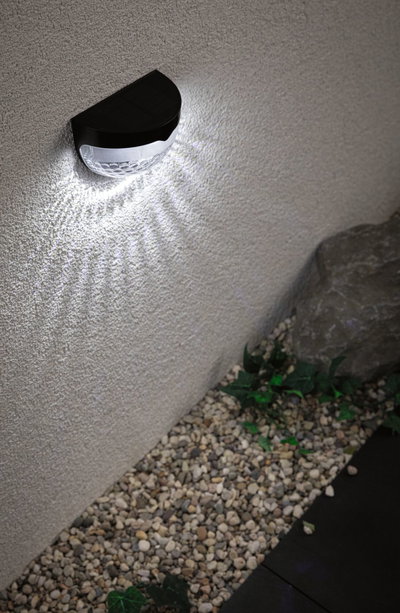 Black solar wall light with white LED on exterior wall above gravel and plants for garden lighting