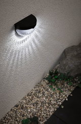 Black solar wall light with white LED on exterior wall above gravel and plants for garden lighting
