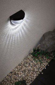 Black plastic solar wall light with white LED illumination on exterior wall above gravel