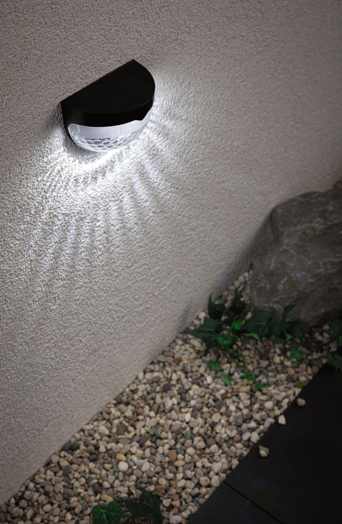 Black plastic solar wall light with white LED illumination on exterior wall above gravel