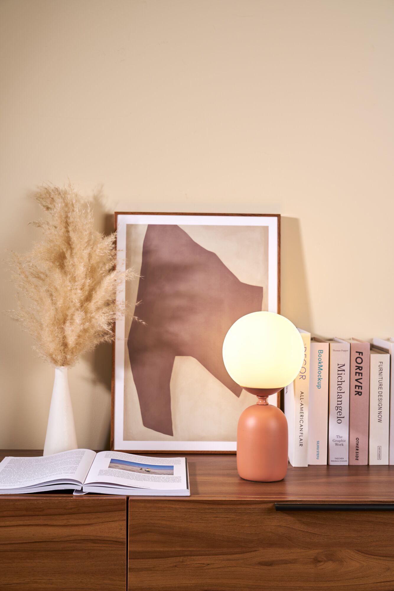 Moderne Tischlampe mit rundem weißem Glas und terrakottafarbenem Keramikfuß auf Holztisch neben Büchern und Kunstbild