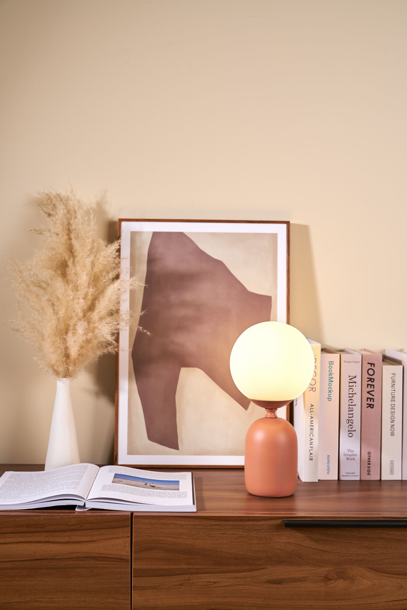 Moderne tafellamp met ronde witte glazen kap en terracotta keramische voet op houten bureau met boeken en ingelijste kunst