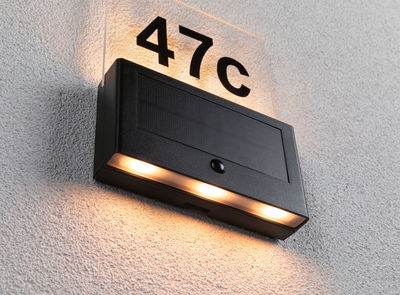 Zwarte zonne-huisnummerlamp met warm wit LED-licht aan witte muur voor buitenverlichting