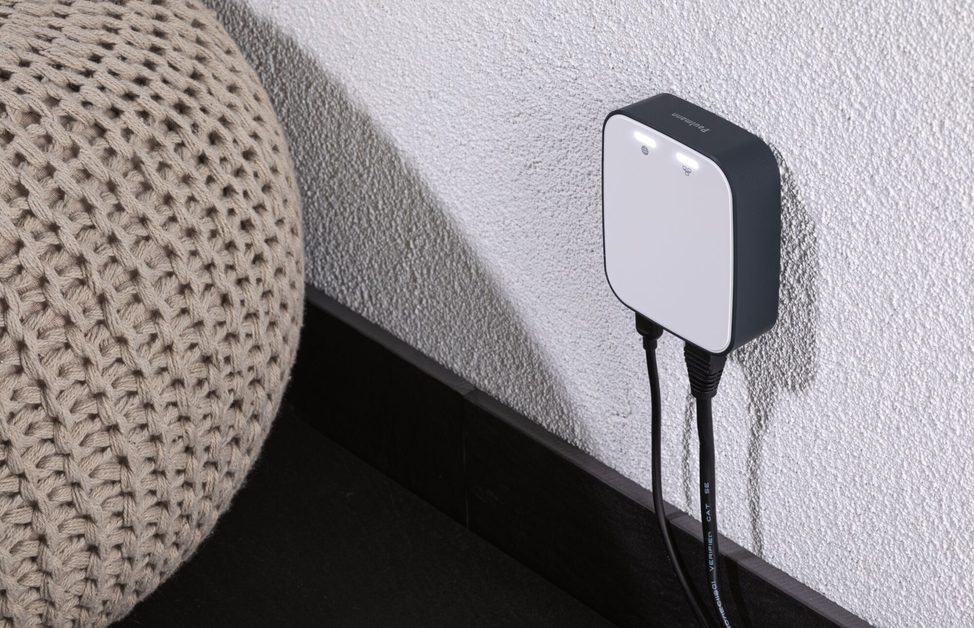 Adaptateur mural compact blanc et noir avec voyants LED sur mur texturé près d’un coussin en tricot beige