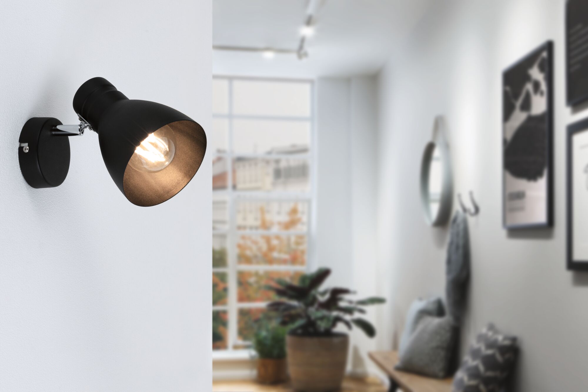 Zwarte metalen wandlamp met verstelbare kap in moderne woonkamer met groot raam en planten.