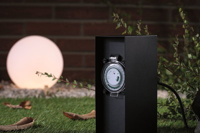 Schwarze Außensteckdose mit integriertem Zeitschaltuhr-Mechanismus im Garten mit beleuchteter Kugelleuchte.