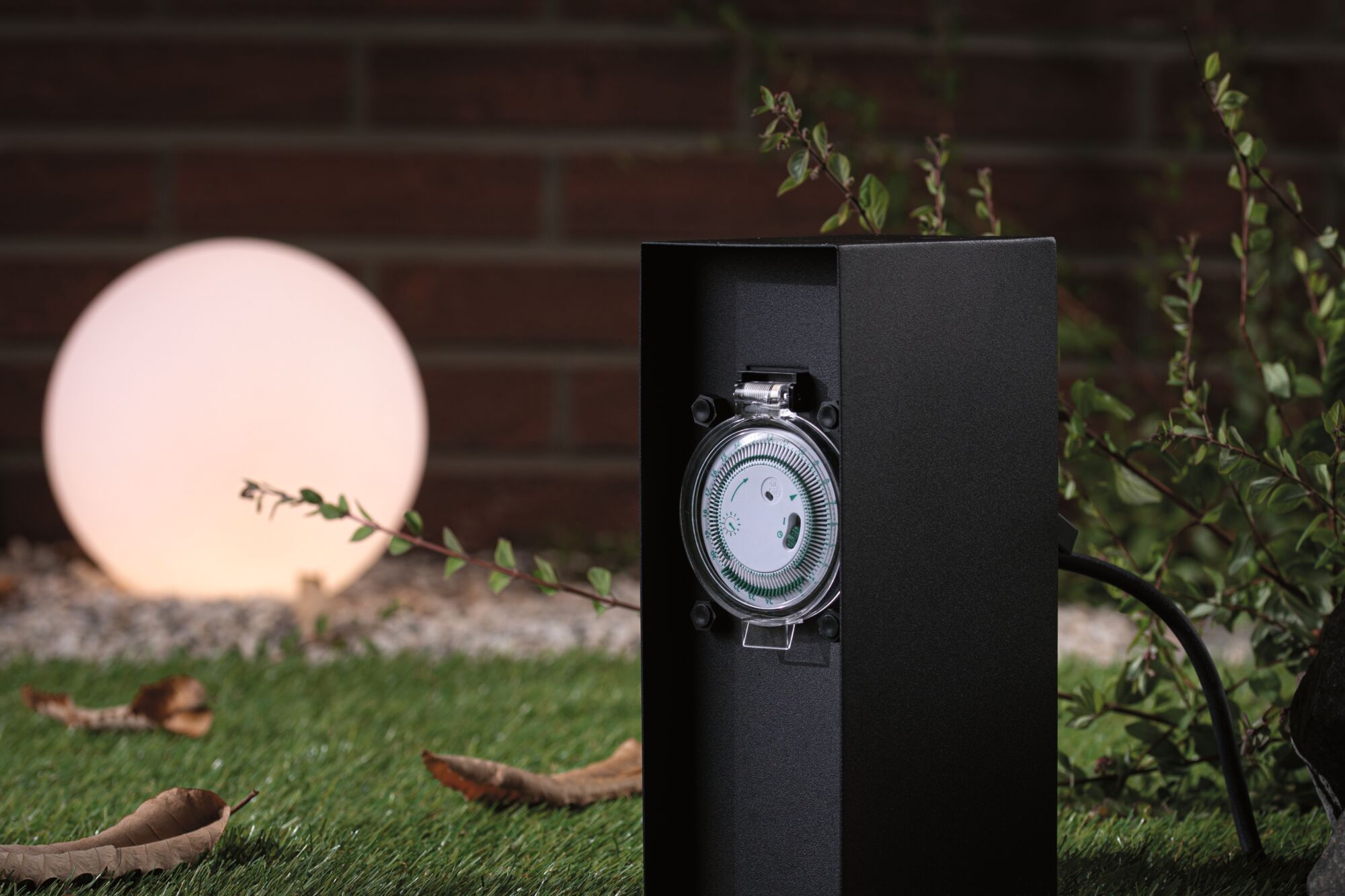Schwarze Außensteckdose mit integriertem Zeitschaltuhr-Mechanismus im Garten mit beleuchteter Kugelleuchte.