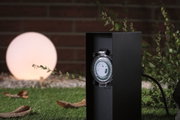 Black outdoor socket with built-in timer mechanism in garden setting with glowing spherical light.