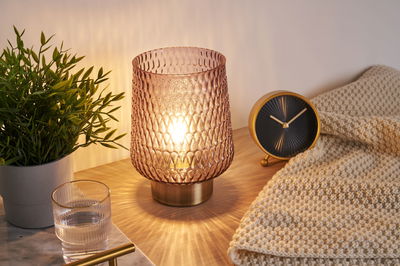 Decorative table lamp with pink textured glass and warm light on wooden surface next to clock and plant