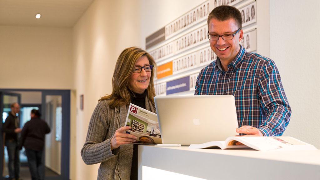 Zwei Kollegen im Büro besprechen Inhalte an einem Laptop und halten Broschüren in der Hand.