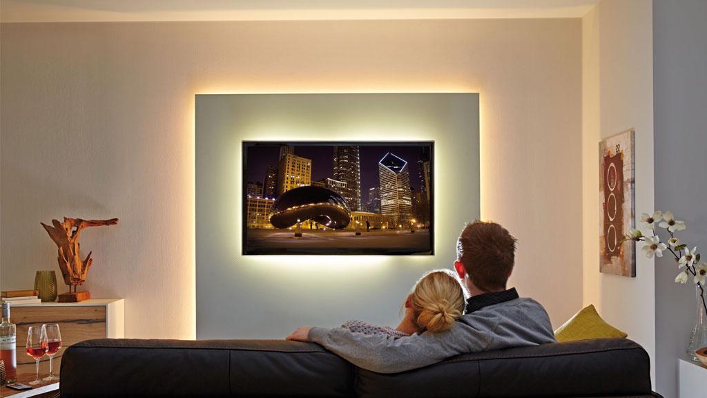 Couple on black sofa watching TV with indirect wall lighting in living room creating cozy ambiance.