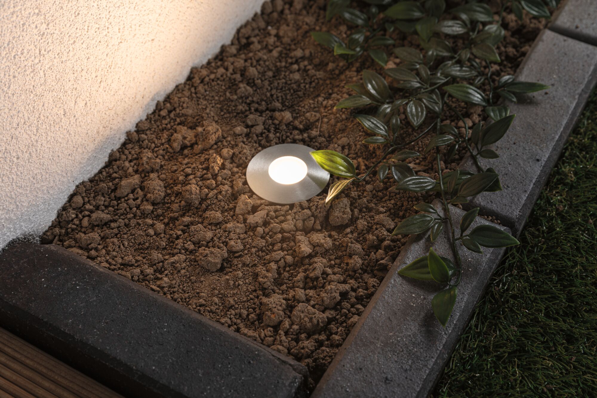 Round stainless steel recessed light with warm white glow in garden bed near green leaves.