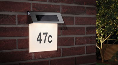 Zonne-energie huisnummerlamp van roestvrij staal met wit verlicht paneel en zwart nummer 47c op rode bakstenen muur