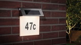 Solar-powered stainless steel house number light with white illuminated panel and black number 47c on red brick wall