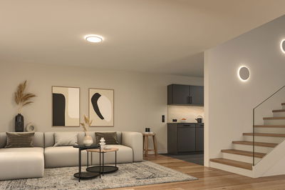 Moderne woonkamer met ronde plafondlamp en wandlampen in warm wit licht, houten trap en grijze bank.