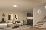 Moderne woonkamer met ronde plafondlamp en wandlampen in warm wit licht, houten trap en grijze bank.