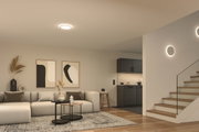 Salon moderne avec plafonnier rond et appliques murales en lumière blanche chaude, escalier en bois et canapé gris.