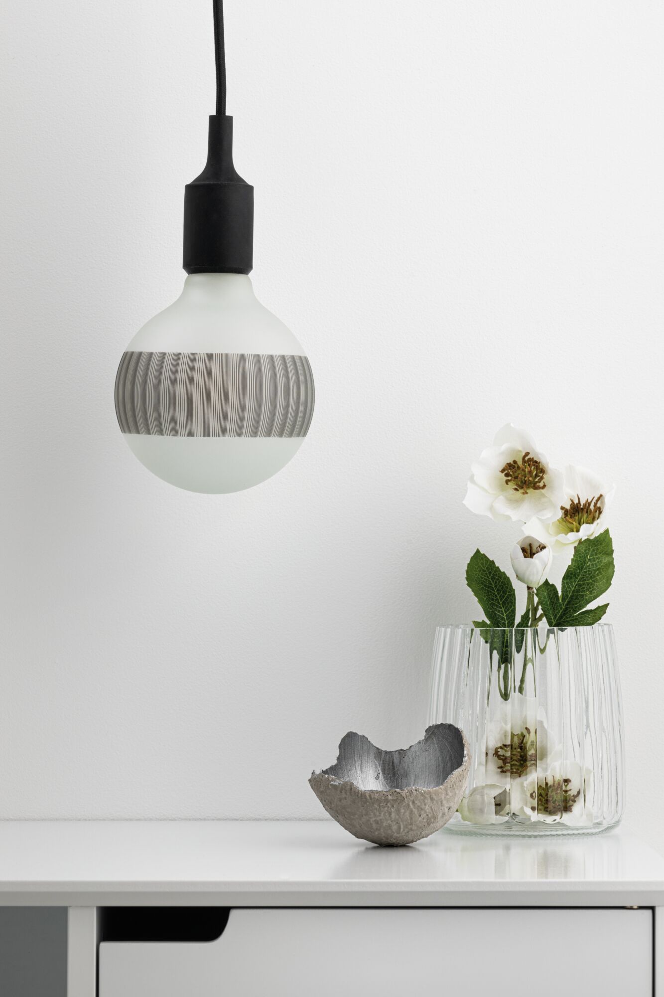 Moderne Pendelleuchte mit mattweißer Glasbirne und schwarzem Kabel über weißem Sideboard mit Vase und Deko.