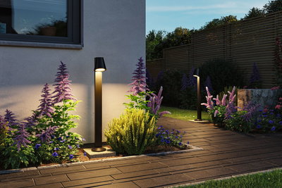 Modern black metal outdoor lights illuminate garden path with colorful flowers and wooden privacy fence.