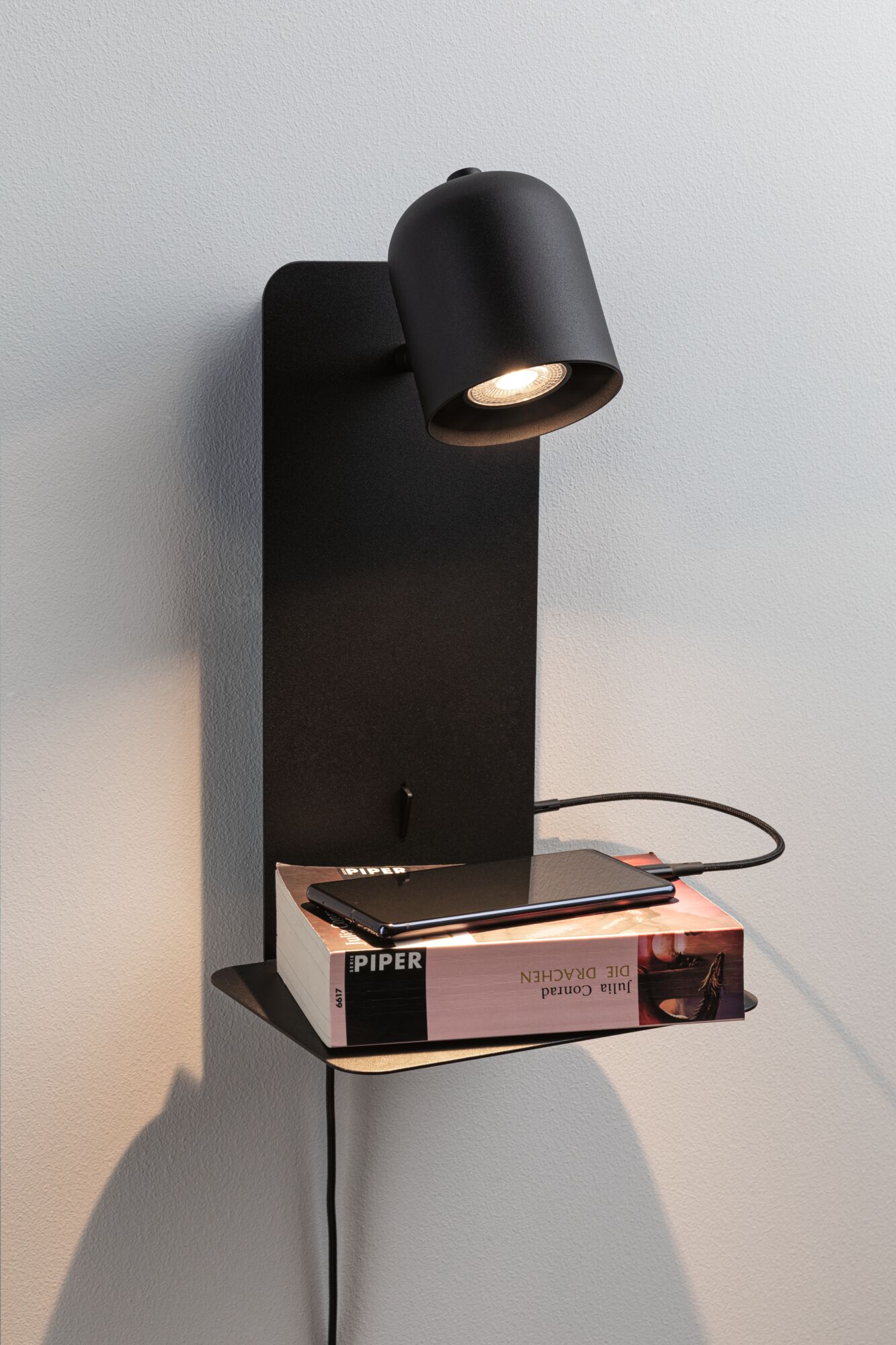 Black metal wall lamp with adjustable spotlight and shelf holding book and smartphone.