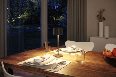 Modern black metal table lamp on wooden dining table with set place and apples, cozy interior lighting.