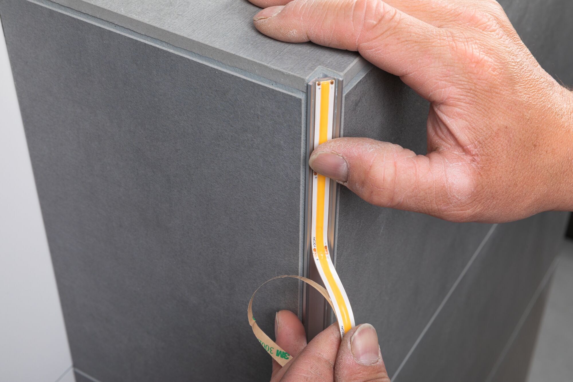 Hand applying self-adhesive LED strip light on grey tile edge for modern interior lighting