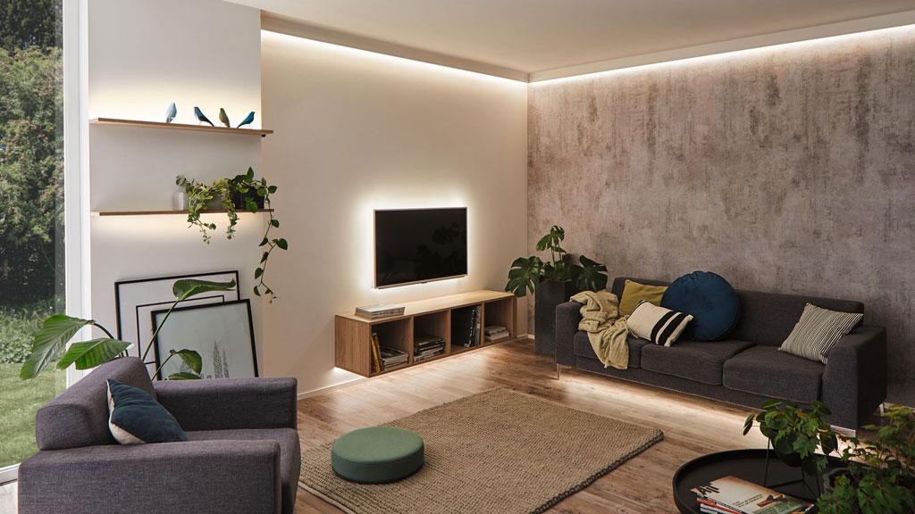 Modernes Wohnzimmer mit indirekter LED-Beleuchtung an Decke und TV-Wand, graue Sofas und Holzregal.