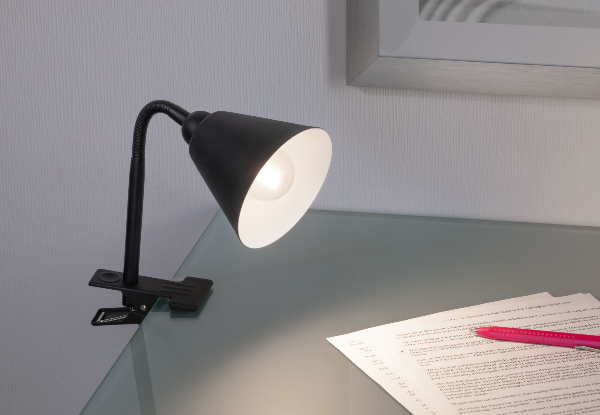 Black clip lamp with flexible neck and white inner reflector on glass desk with documents and pen.