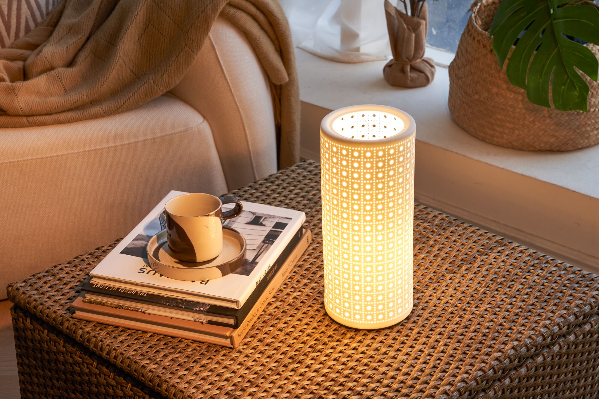 White ceramic cylindrical lamp with perforated pattern on wicker table near sofa and cup