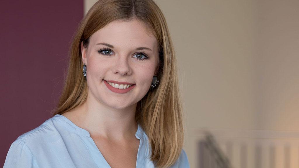 Junge Frau mit blauer Bluse und Ohrringen vor neutralem Hintergrund, freundliches Porträtfoto