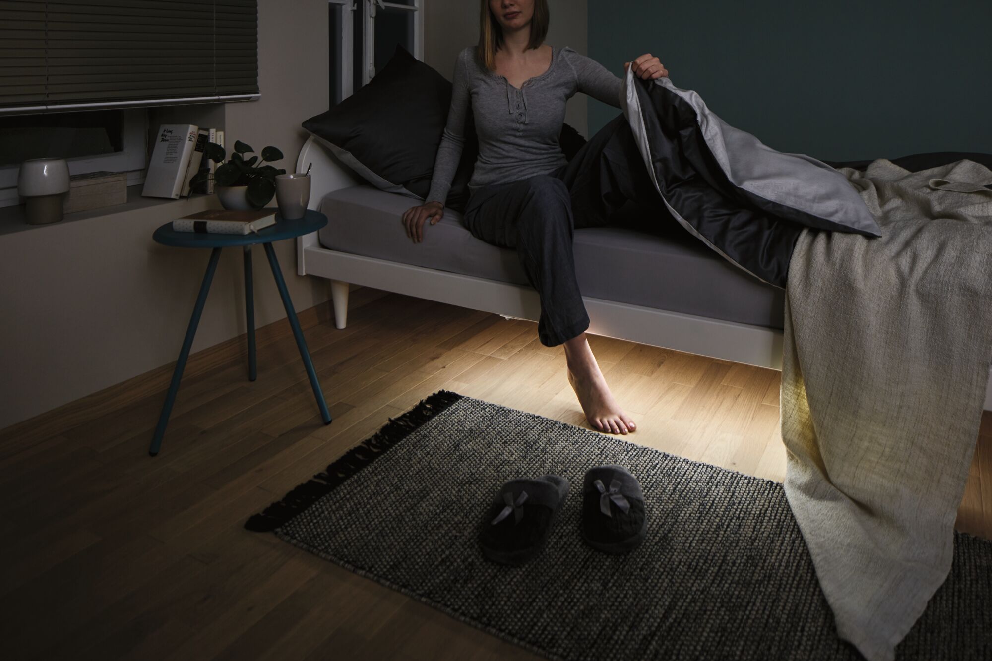 Dark bedroom with grey bed, black blanket, and slippers on rug over wooden floor at night.