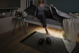 Dark bedroom with grey bed, black blanket, and slippers on rug over wooden floor at night.