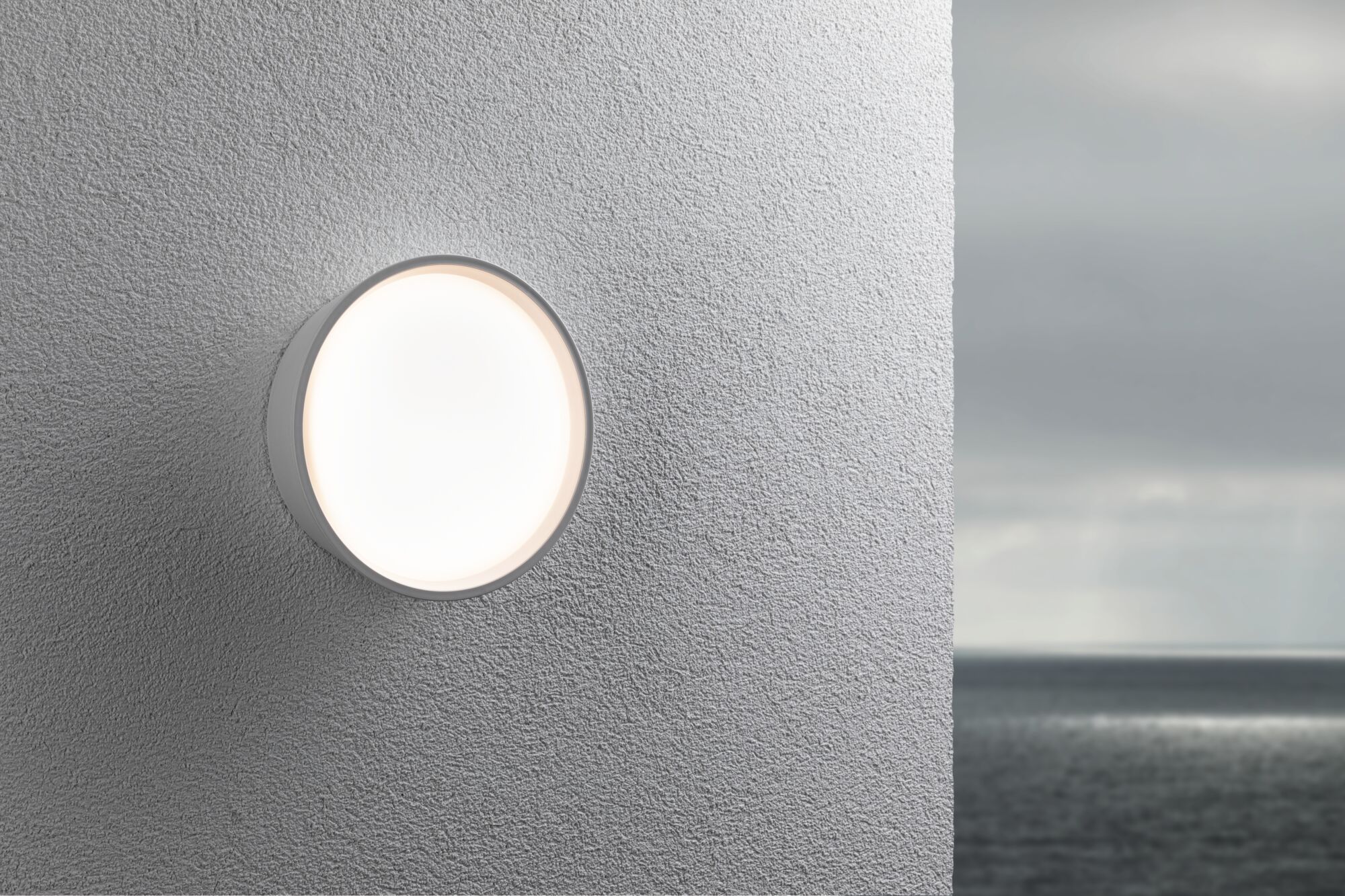 Runde weiße Außenwandleuchte aus Kunststoff an grauer Wand mit Meereshintergrund, LED Licht