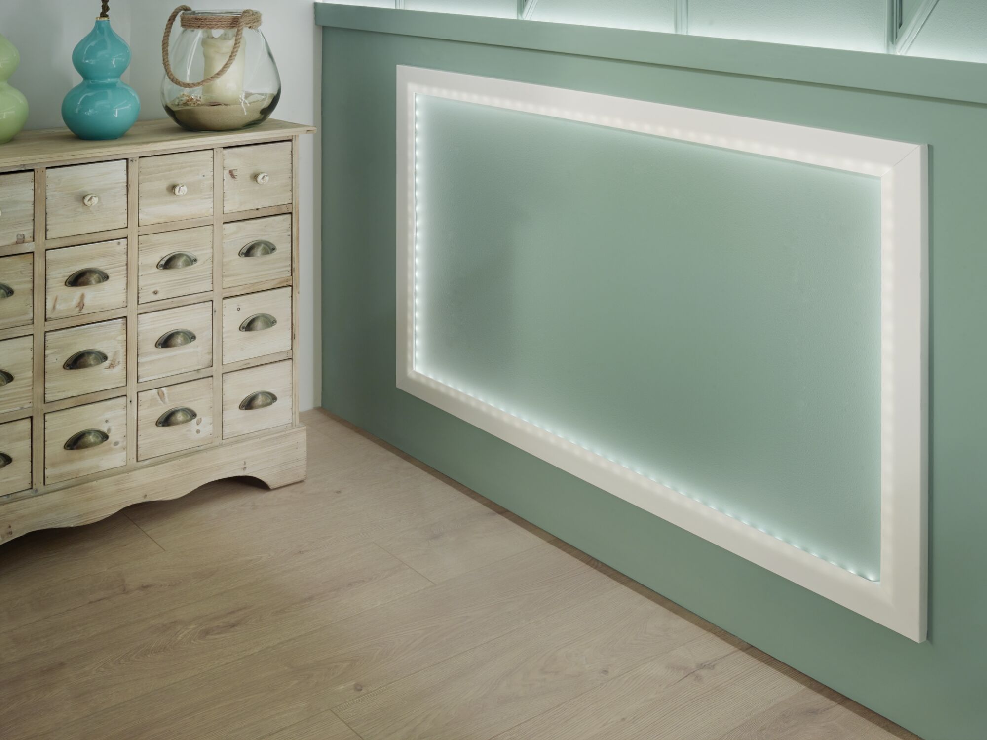 Weiße LED-Rahmenleuchte an grüner Wand mit Holzkommode und Deko im modernen Wohnraum