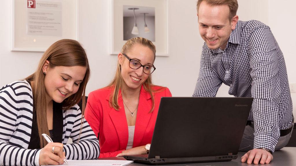 Drei Personen in Büro beraten sich gemeinsam an einem schwarzen Lenovo Laptop, Arbeitsumgebung.