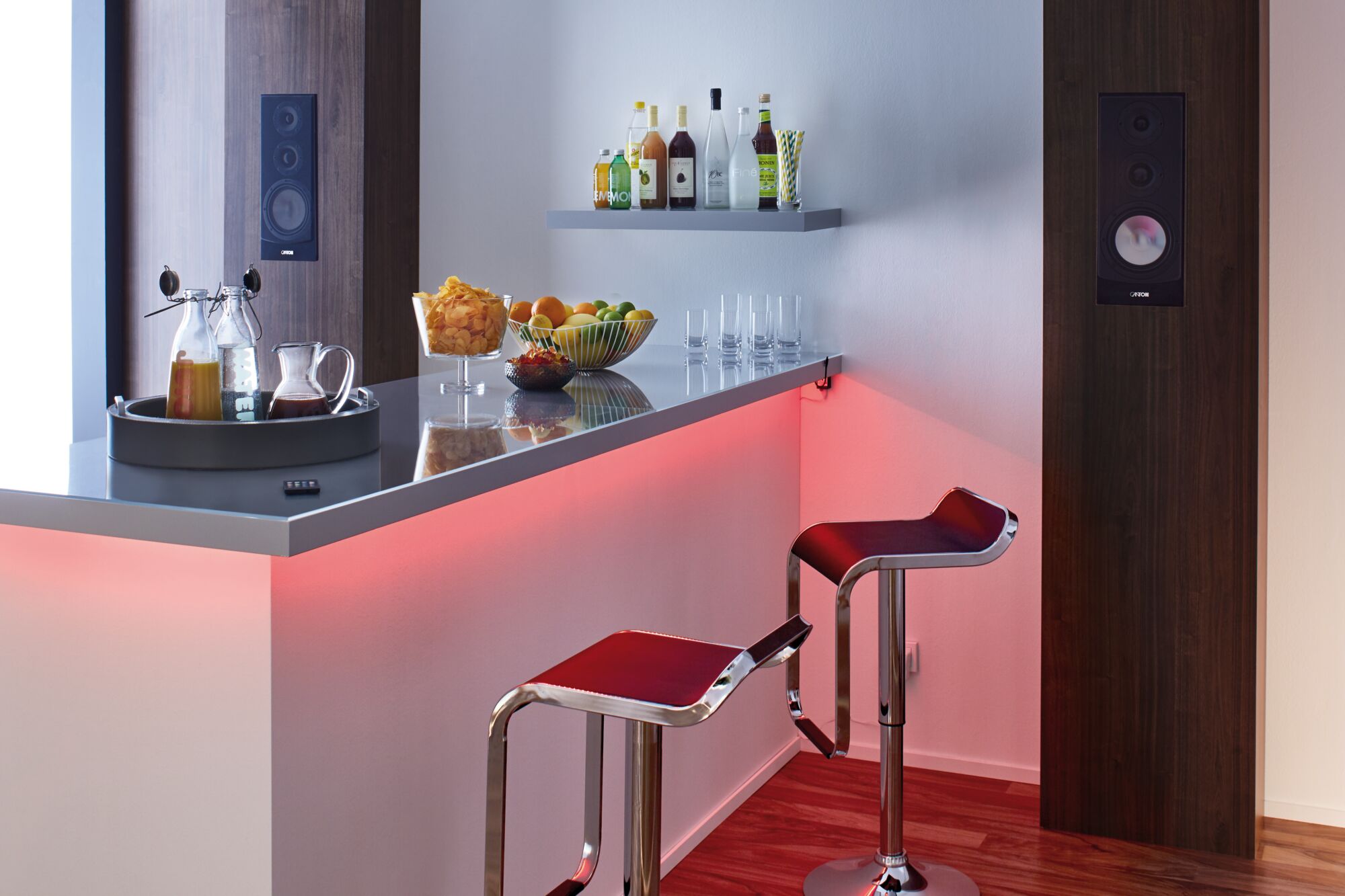 Modern bar area with red LED lighting, two black bar stools, and wooden wall speakers in a lounge setting