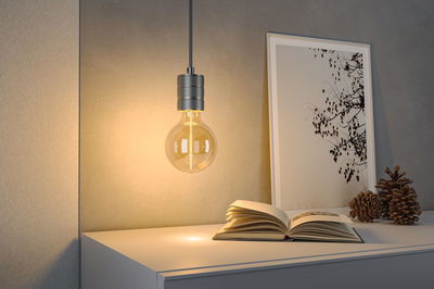 Hanging light bulb with warm white glow above white table with open book and decor.