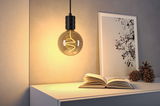 Hängende Glühbirne mit warmweißem LED-Filament über weißem Tisch mit Buch und Deko im Wohnzimmer