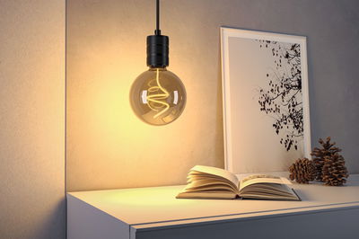 Hängende Glühbirne mit warmweißem LED-Filament über weißem Tisch mit Buch und Deko im Wohnzimmer