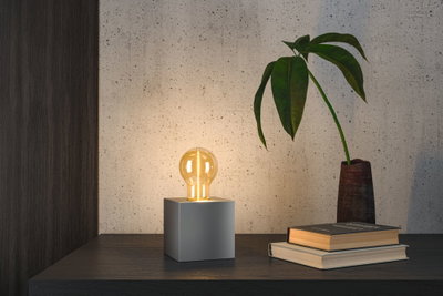 Modern table lamp with grey metal cube and warm white LED bulb on dark wooden surface.