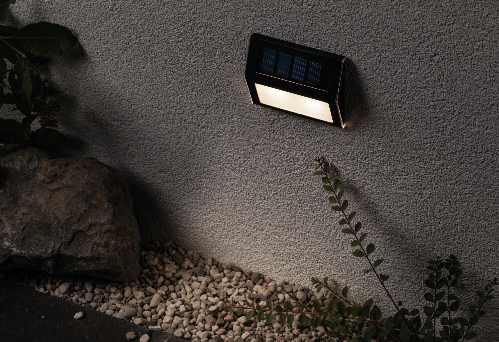 Schwarze Solar-Wandleuchte mit warmweißem LED-Licht an grauer Außenwand über Kies und Pflanzen