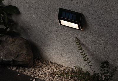 Schwarze Solar-Wandleuchte mit warmweißem LED-Licht an grauer Außenwand über Kies und Pflanzen